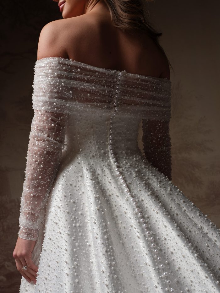 Maggie Sottero Elowyn Bridal Jacket, back view detail of the pearl and sequin-encrusted off-the-shoulder jacket with sheer long sleeves, worn over a matching pearl-covered ballgown skirt.