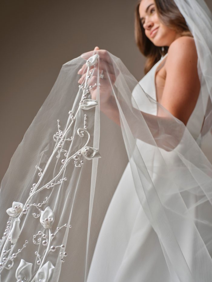 
Maggie Sottero Lawrence Veil Accessory, close-up detail of the veil's embellished edge showing 3D calla lily appliqués, delicate scrolling vine embroidery, and scattered pearl beading along the tulle border.