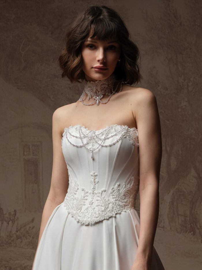 Close-up of the Seattle Wedding Dress Choker by Maggie Sottero paired with a coordinating wedding gown. The accessory features a lace choker embellished with pearls, crystals, and draped beaded strands that frame the neckline. The gown showcases a strapless corset bodice with exposed boning-inspired seaming, delicate lace appliqués, and beaded detailing along the neckline and waist, creating a romantic, Victorian-inspired bridal look.