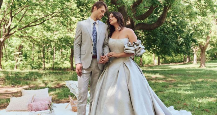 Bride wearing the plus-size Fortune gown by Maggie Sottero, a strapless ball gown with dramatic off-shoulder ruffle sleeve, holding hands with groom in a wooded garden setting.