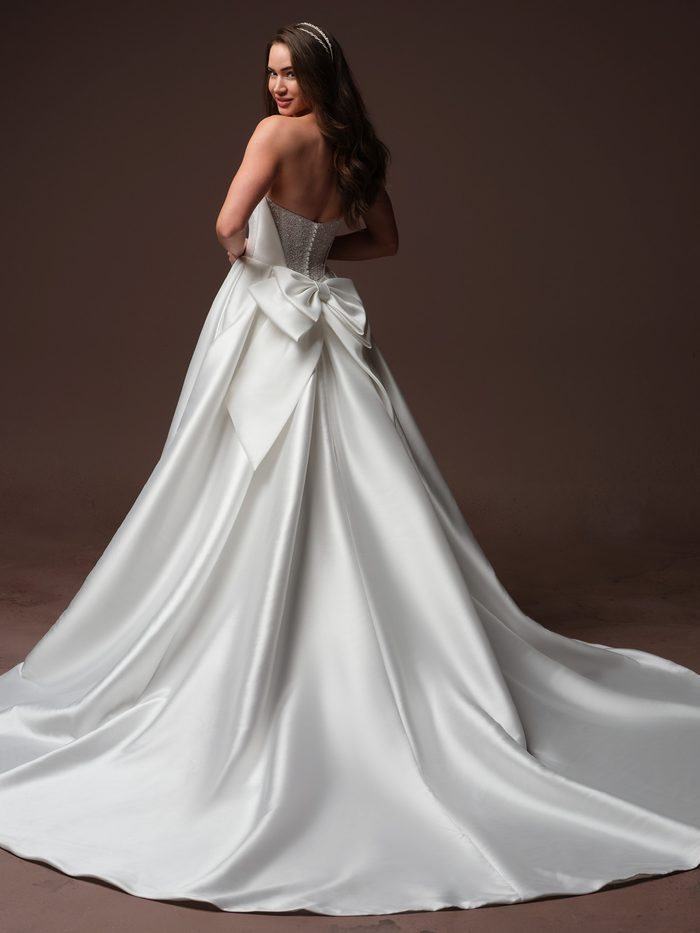 Back view of the Joanie by Rebecca Ingram wedding gown featuring a strapless satin ballgown silhouette with a low back, fitted bodice, and dramatic cathedral-length train. A statement oversized satin bow accents the back waist, creating a striking focal point while adding volume and romance to the embelished design.