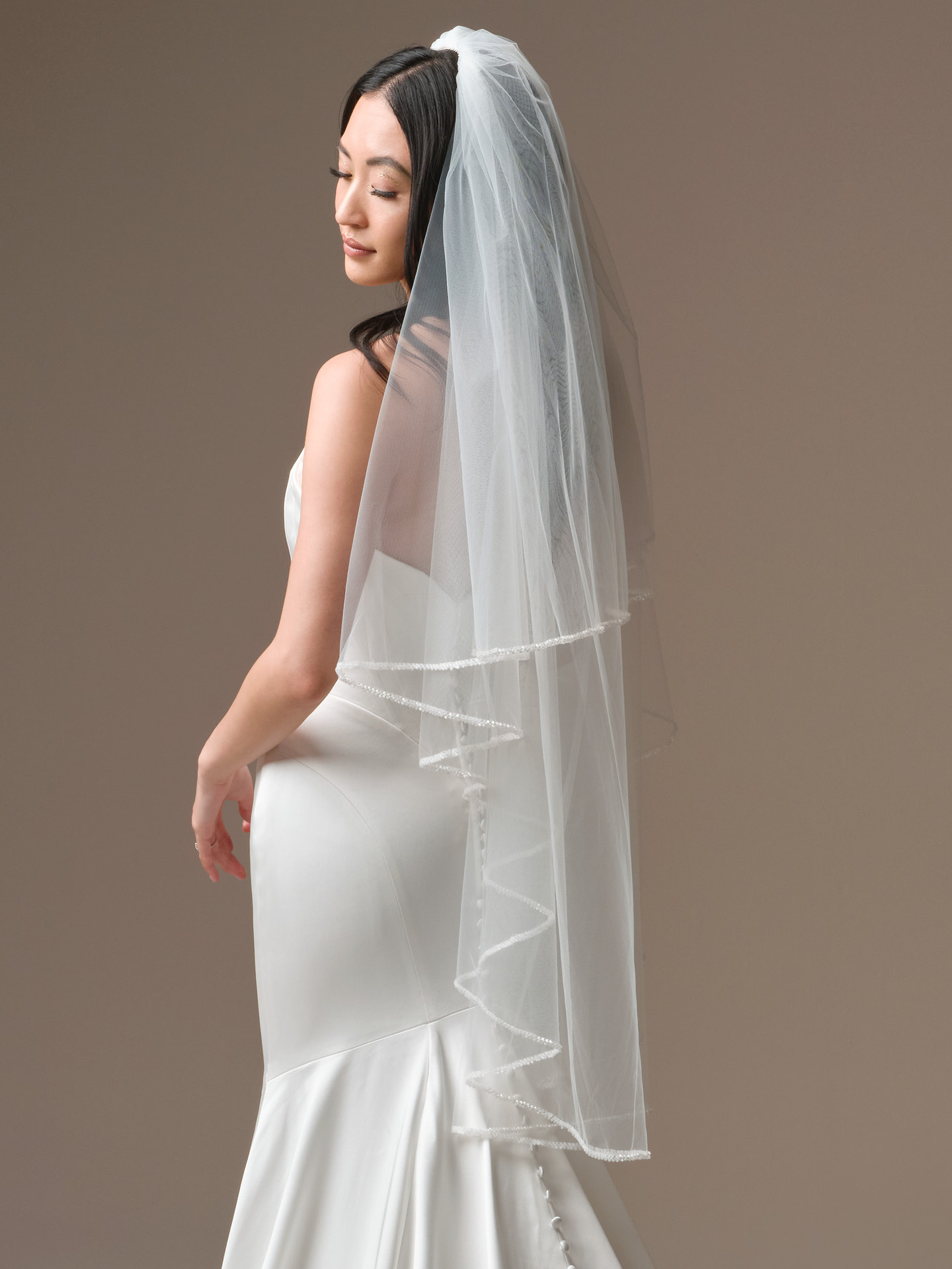 Bride wearing a fingertip veil from the Maggie Sottero Essentials Edit featuring delicate beaded trim along the edge, styled over a fitted satin wedding dress with a soft flared hem.