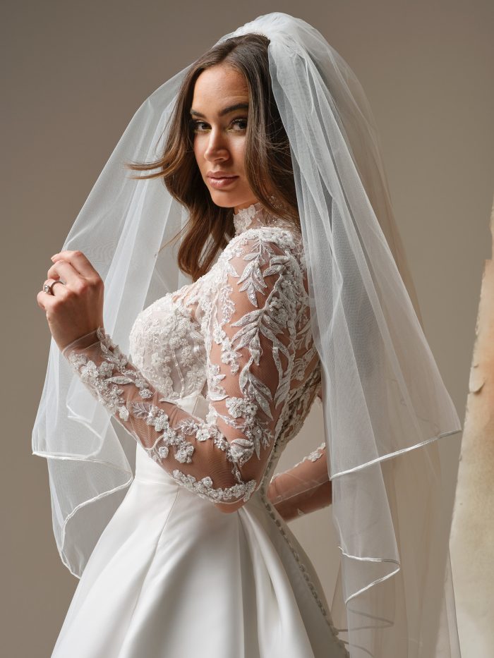 Bride wearing a soutache edge veil from the Maggie Sottero Essentials Edit, featuring a delicate corded trim along the edge, styled over a long-sleeve lace wedding gown with a full satin skirt.