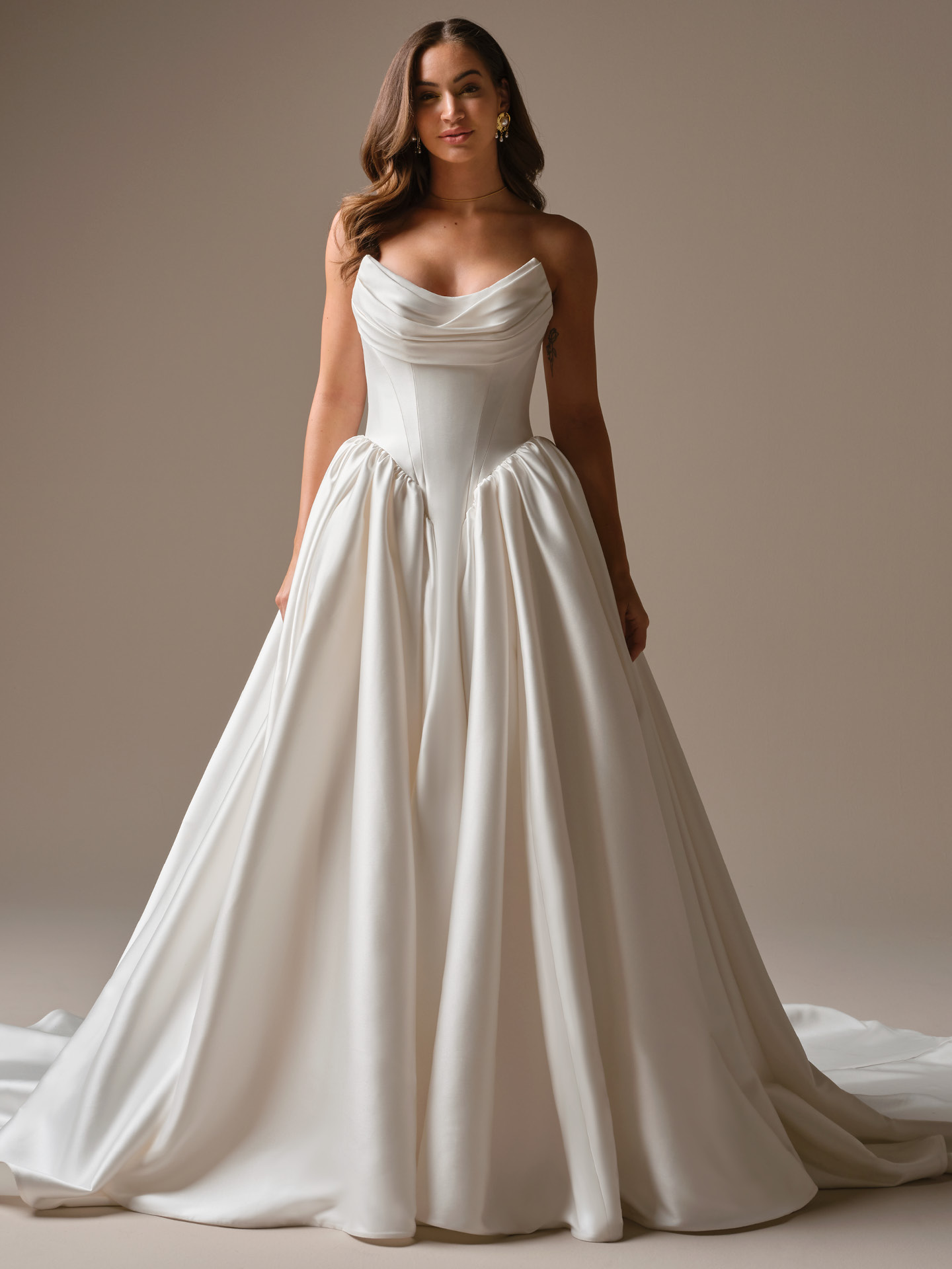 Bride wearing Hudson by Maggie Sottero, a strapless satin ballgown wedding dress featuring a softly draped cowl neckline, structured corset bodice, gathered skirt panels, and a sweeping train.