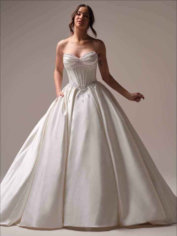 Full-length view of the Juneau ballgown satin wedding dress by Maggie Sottero featuring a strapless sweetheart neckline, pearl-accented corset bodice, and a dramatic satin skirt with pockets.