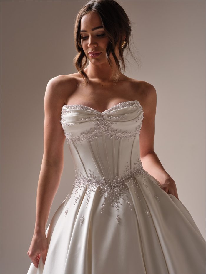 Close-up of the Juneau satin wedding dress by Maggie Sottero showcasing the ruched sweetheart neckline, structured corset detailing, and intricate pearl embellishments across the bodice and waist.