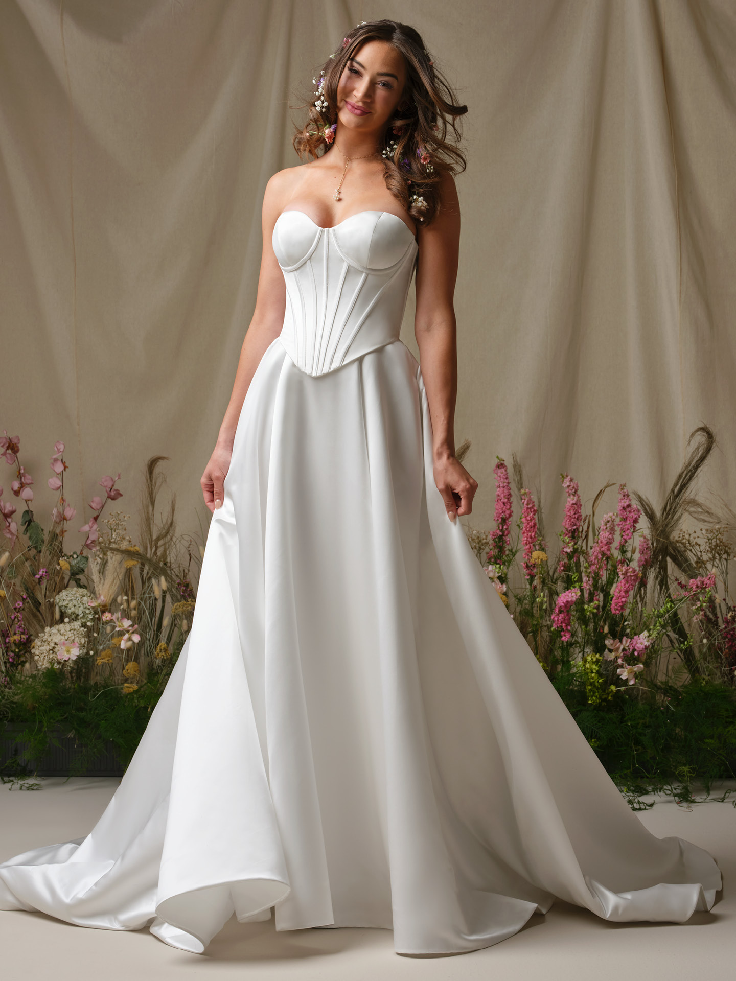 Bride wearing Alexia by Rebecca Ingram, a strapless satin A-line wedding dress featuring a structured corset bodice with defined cups and a full flowing skirt, styled in a romantic floral setting.