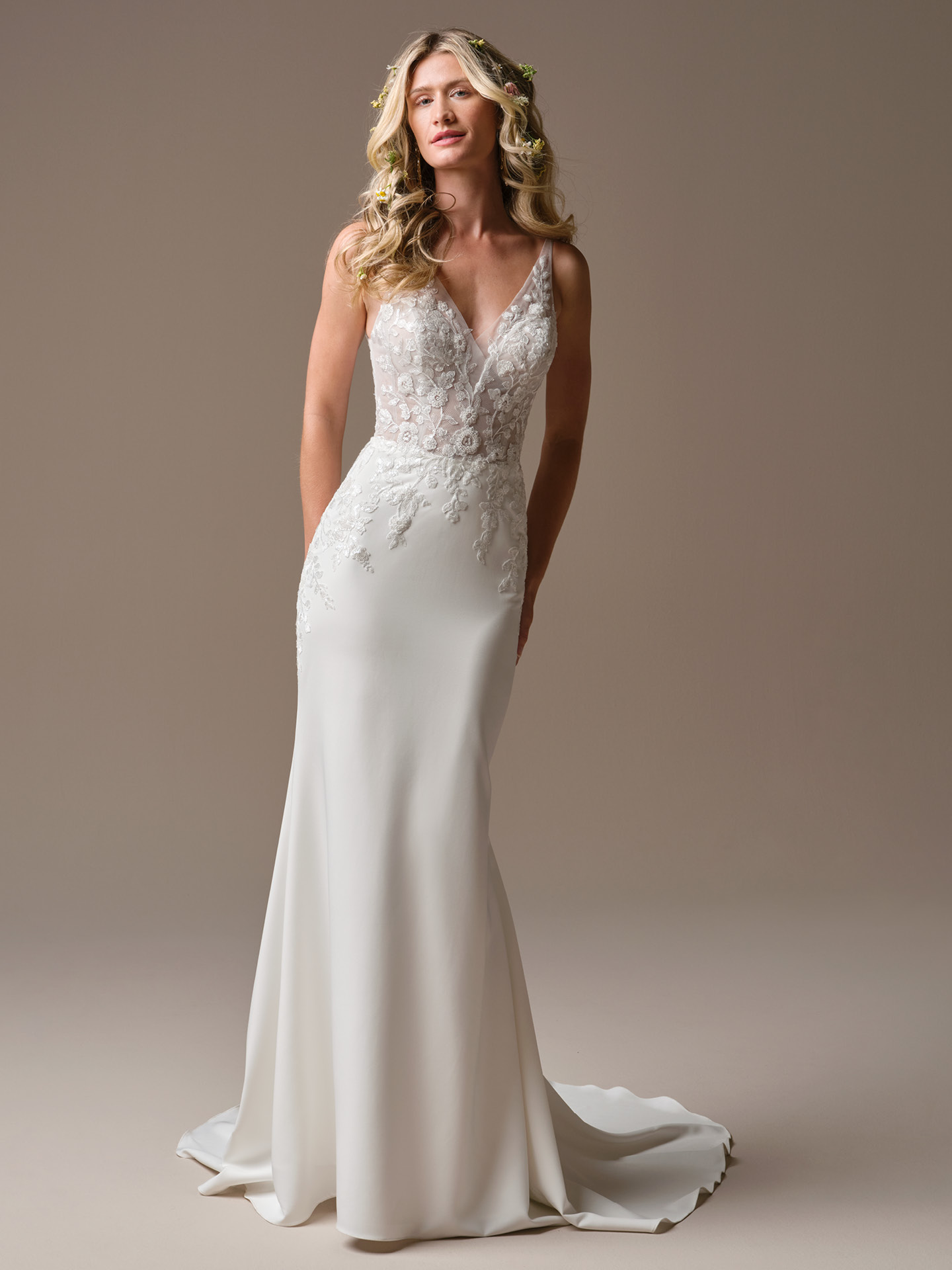 Bride wearing Holly by Rebecca Ingram, a sleeveless sheath wedding dress featuring a deep illusion V-neckline, lace appliqué bodice, smooth fitted skirt, and a subtle chapel-length train.