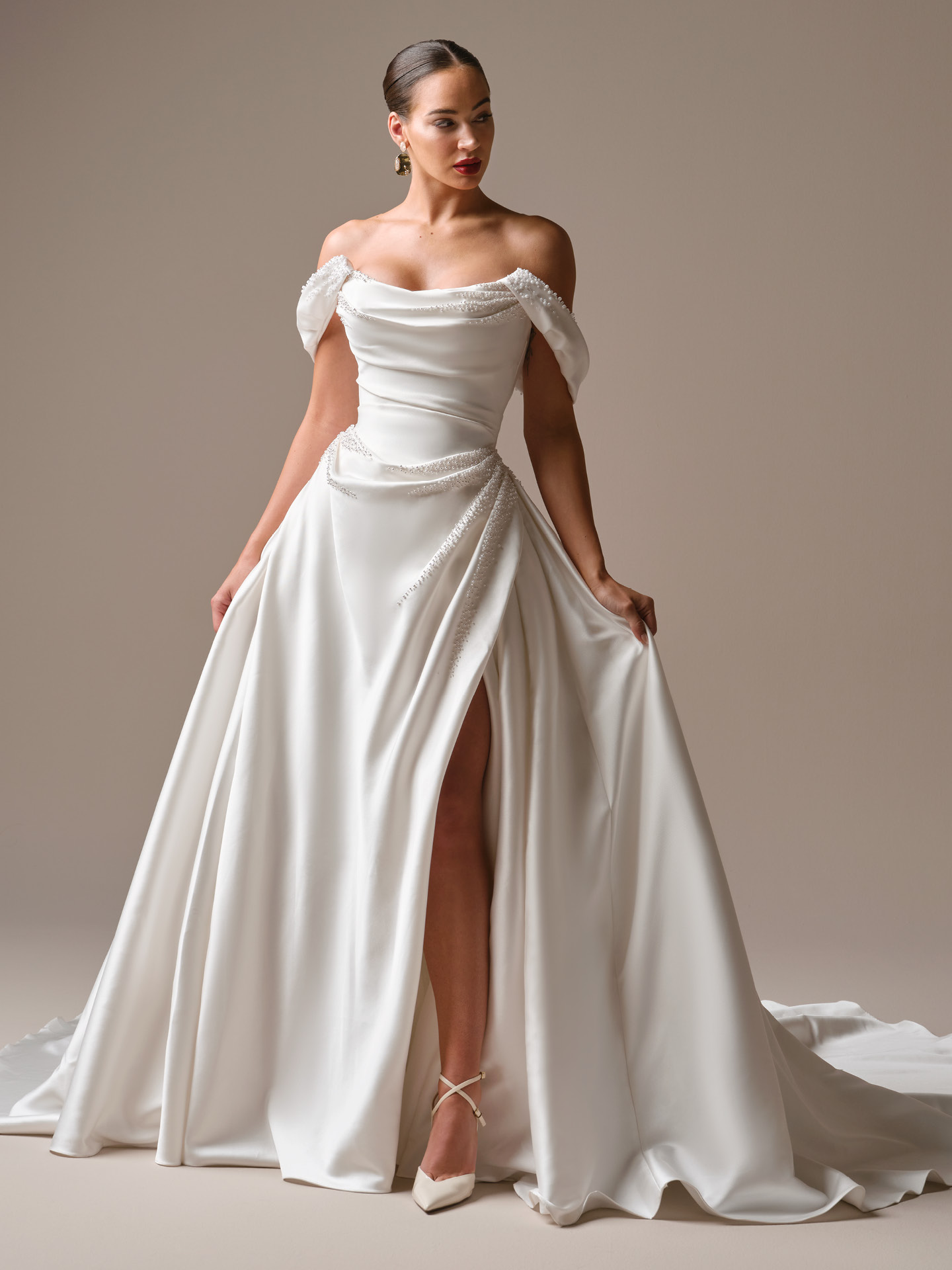 Bride wearing Kincaid by Sottero and Midgley, an off-the-shoulder satin ballgown wedding dress with draped bodice, pearl-accented detailing, asymmetrical waist embellishment, thigh-high slit, and a full flowing skirt with a sweeping train.