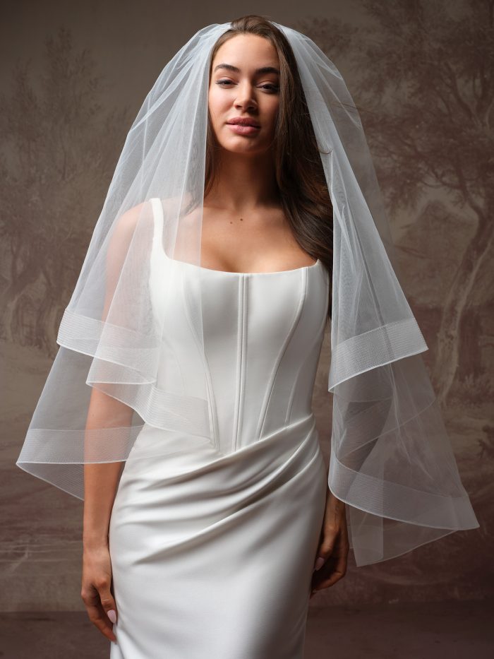 Front view of the Bellissima by Maggie Sottero elbow-length wedding veil from the Essentials Edit collection, a two-tier plain-edge tulle veil styled over a sleeveless square-neck satin fitted gown.