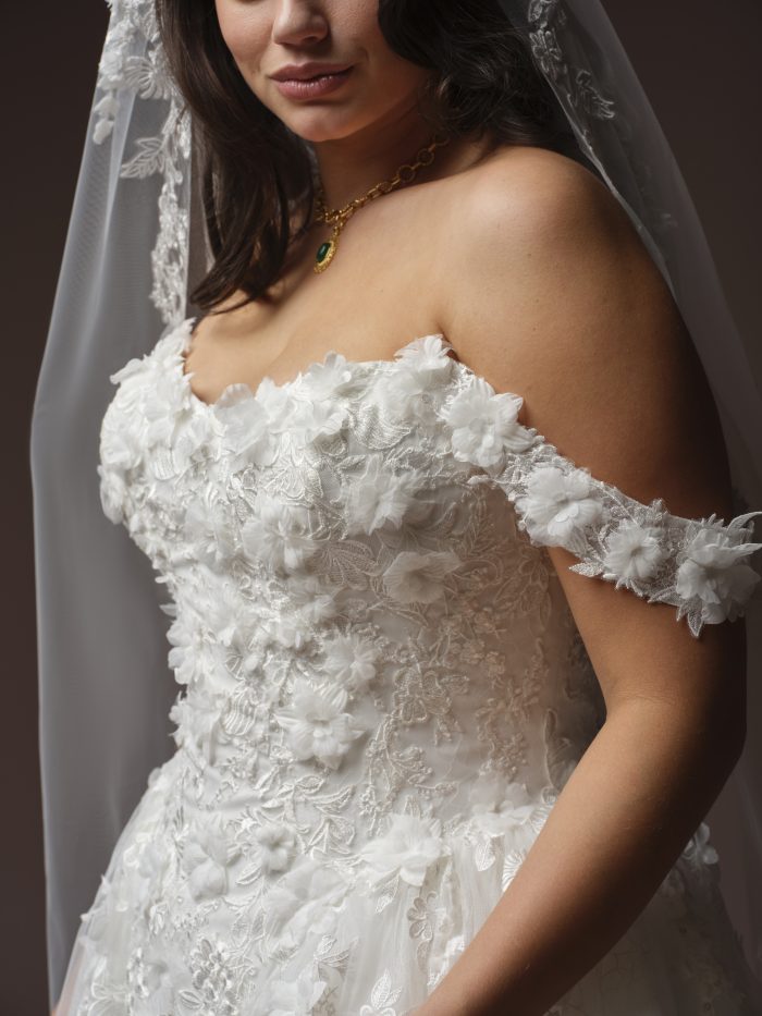 Benton ballgown wedding dress by Maggie Sottero with an off-the-shoulder bodice, cap sleeves, and dimensional floral appliqués, shown in a close-up highlighting textured lace and floral details.