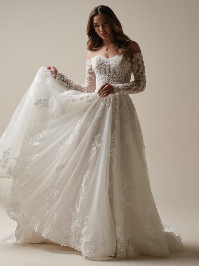 Rita A-line wedding dress by Maggie Sottero featuring off-the-shoulder lace sleeves, lace bodice, and flowing tulle skirt.