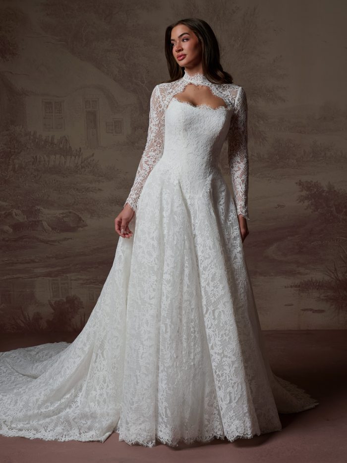 Serena lace jacket by Maggie Sottero featuring a high neckline, long sleeves, and scalloped lace edging, shown over a strapless lace A-line wedding dress in a full-length view.