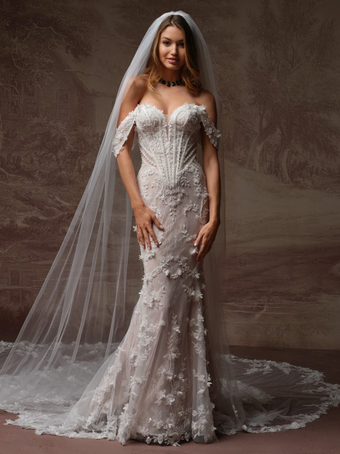 Stasia wedding dress by Maggie Sottero with a fitted silhouette, illusion bodice, and dimensional floral lace, paired with cap sleeves and a long sheer veil, shown from the front.