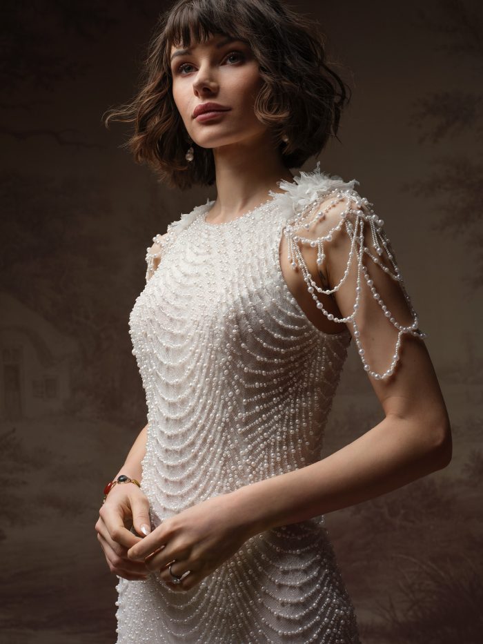 Oleta sheath wedding dress by Sottero and Midgley featuring an allover beaded design with a fitted silhouette and beaded caplet sleeves draped in strands, shown in a close-up view.
