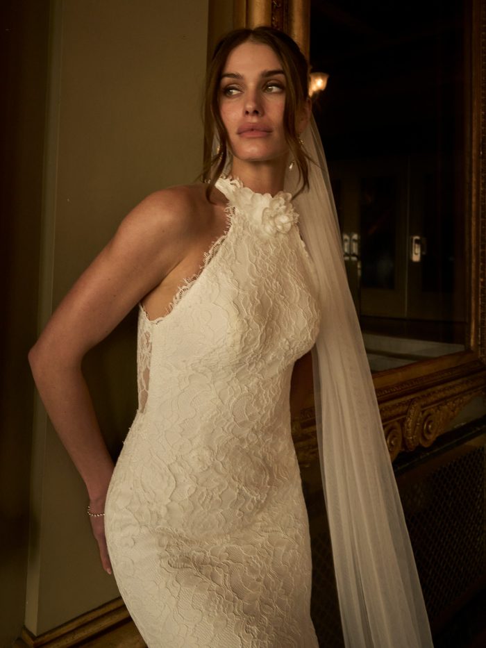 Anya sheath wedding dress by Maggie Sottero in a close-up view, highlighting the intricate lace texture, halter neckline with floral accent, and scalloped edges along the bodice. The look is completed with a sheer veil in a softly lit, classic interior setting.