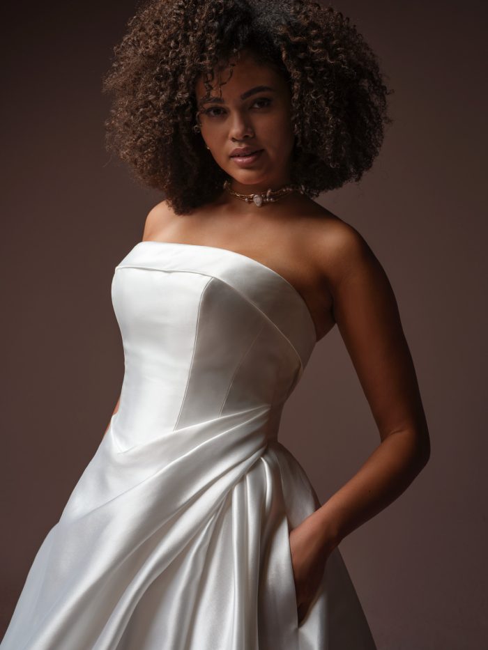 Rebecca Ingram Agatha A-line wedding dress, close-up detail — strapless satin bodice with clean seaming and draped asymmetrical skirt with pocket detail.