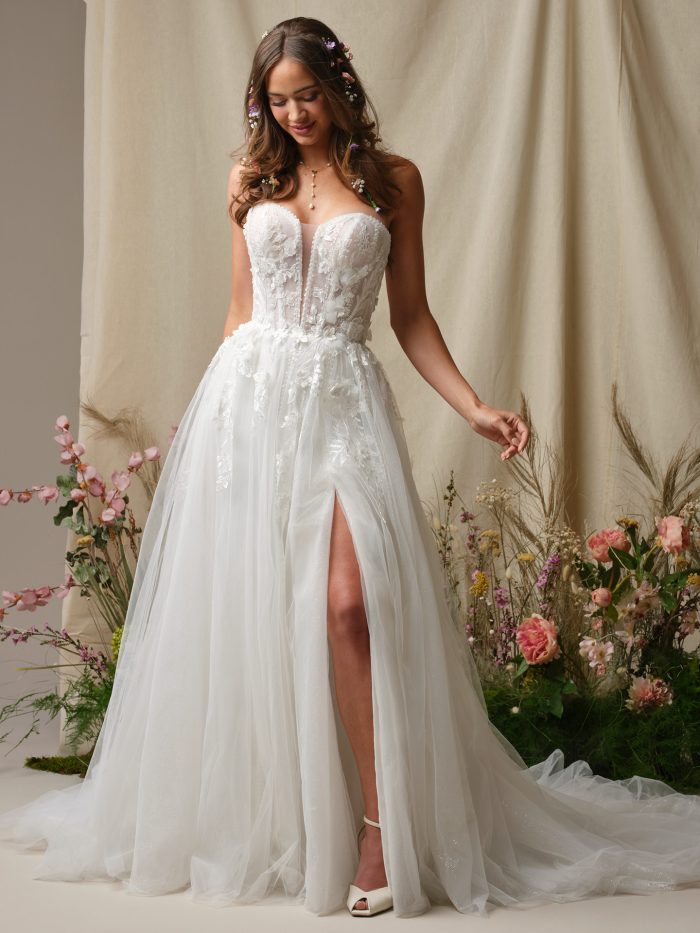 Nora by Rebecca Ingram strapless A-line wedding dress with 3-D floral lace, illusion plunge neckline, and flowing tulle skirt with side slit