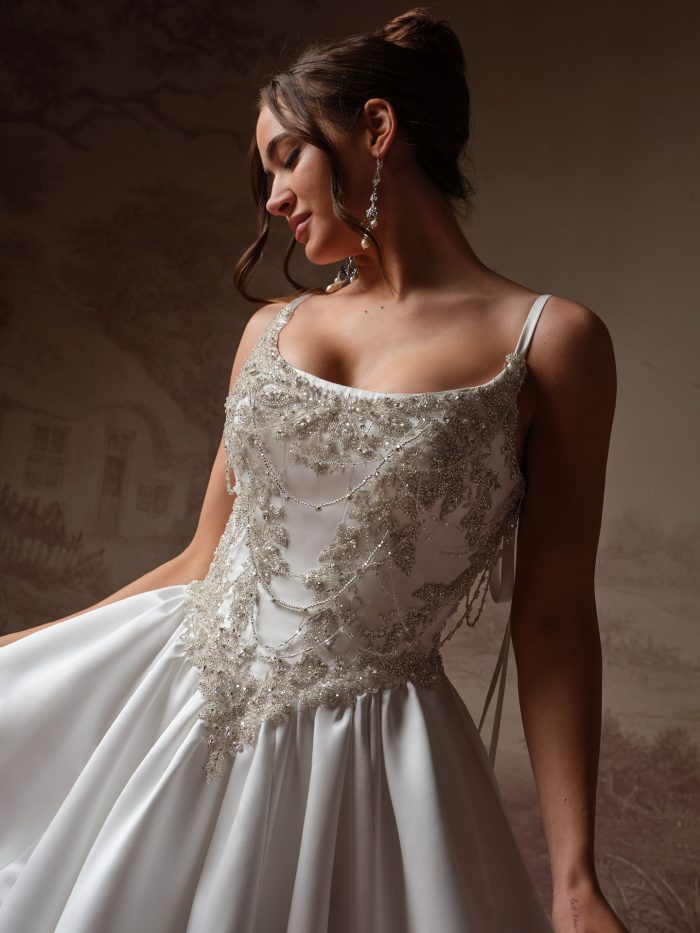 Sottero and Midgley Aelarion A-line wedding dress, close-up detail — beaded lace bodice with draped chain embellishment and thin straps over satin skirt.