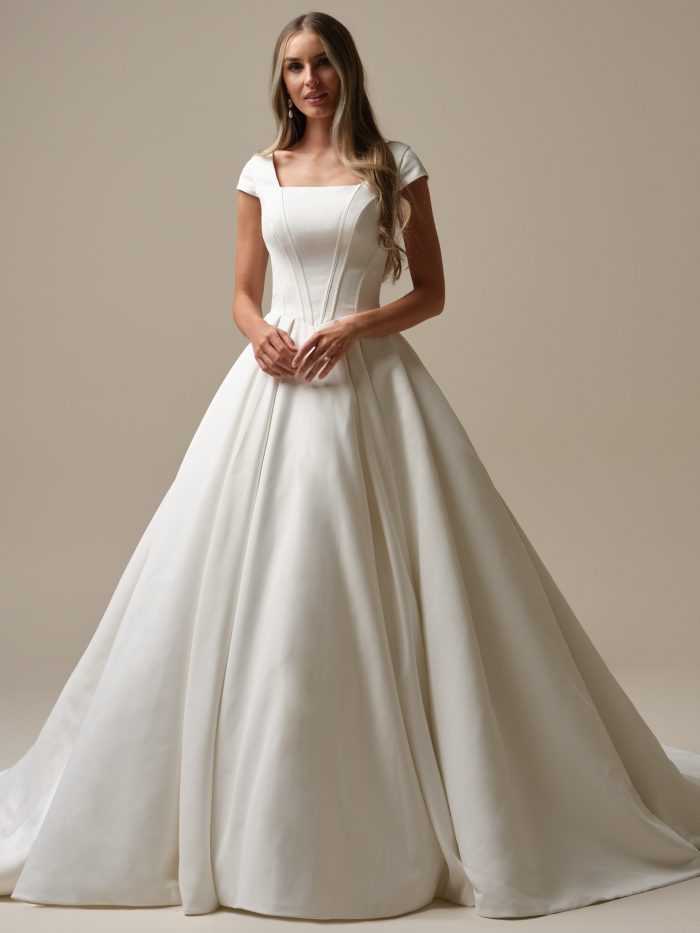 Full-length front view of Derrick Leigh by Maggie Sottero a satin ballgown wedding dress featuring a structured bodice with corset-style seam detailing, a straight neckline with cap sleeves, and a voluminous pleated skirt that extends into a dramatic train, photographed against a neutral studio background.