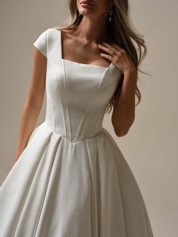 Close-up detail of Derrick Leigh by Maggie Sottero a ballgown wedding dress bodice made from smooth satin, featuring a square neckline, wide cap sleeves, and visible corset-style seam detailing, paired with a softly pleated skirt, photographed against a neutral studio background.