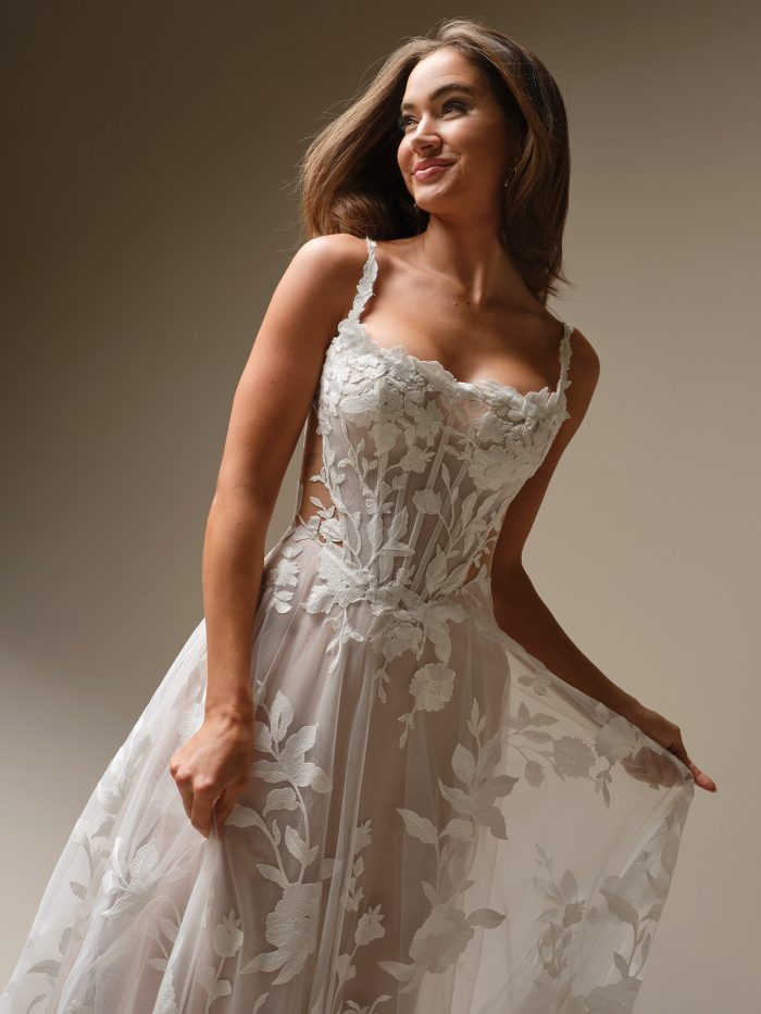 Close-up view of Natalie Marie by Maggie Sottero an A-line wedding dress featuring a sheer, floral lace bodice with visible corset structure, delicate lace straps, and a flowing tulle skirt with large-scale lace appliqués, photographed against a neutral studio background.