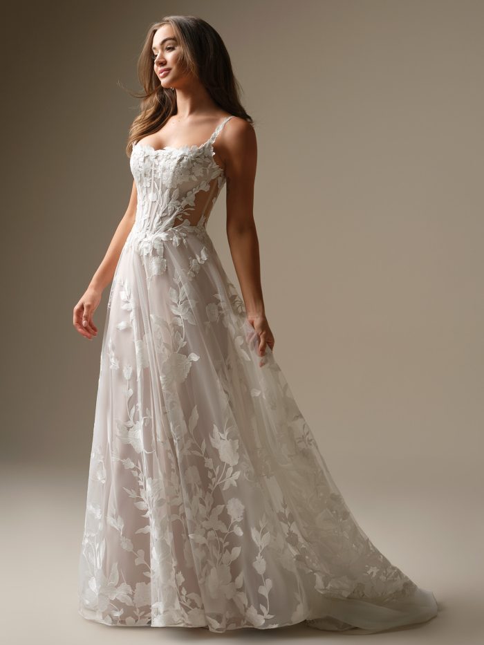 Three-quarter view of Natalie Marie by Maggie Sottero an A-line wedding dress with a sheer, floral lace corset-style bodice, delicate lace straps, and a softly flowing tulle skirt with large floral appliqués, photographed in a neutral studio setting.