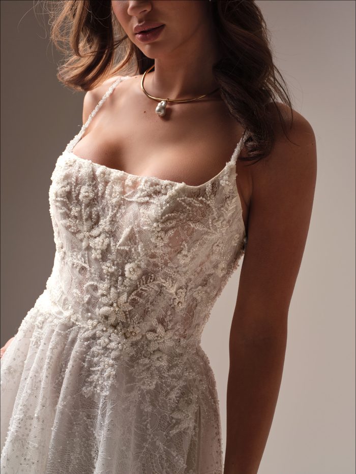 Close-up of Salvadora by Maggie Sottero an A-line wedding dress featuring delicate lace with floral beadwork, thin spaghetti straps, and a softly structured bodice, paired with a gold choker necklace with a pearl pendant against a neutral studio background.