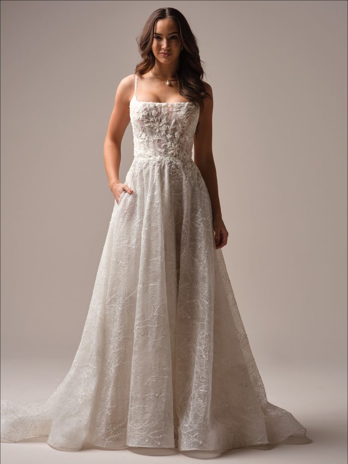 Full-length view of Salvadora by Maggie Sottero an A-line wedding dress with a fitted lace-and-beaded bodice, thin spaghetti straps, and a flowing lace skirt with subtle sparkle, shown on a model standing against a neutral studio background.