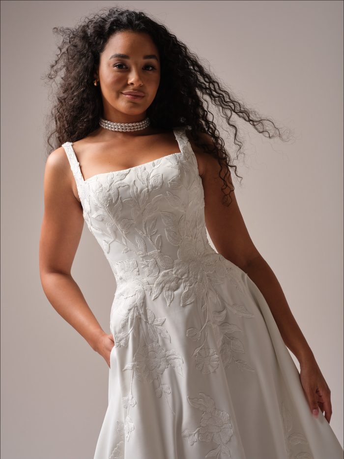 Close-up of Sheridan by Maggie Sottero an A-line wedding dress bodice featuring smooth satin fabric with tonal floral appliqué embroidery, wide shoulder straps, and a softly angled neckline, styled with a pearl choker necklace and photographed against a neutral studio background.