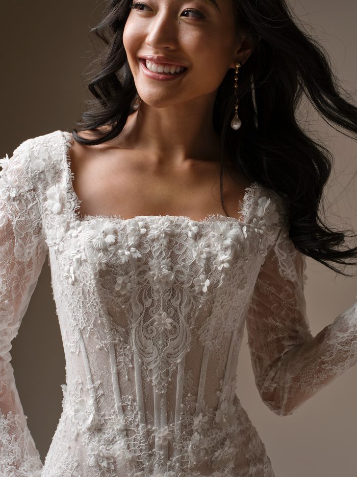 Close-up detail of Takera Leigh by Maggie Sottero a fit-and-flare wedding dress bodice featuring sheer structured lace with three-dimensional floral appliqués, a straight neckline, and long illusion lace sleeves, photographed against a neutral studio background.