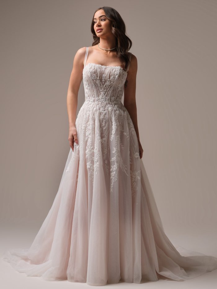 Full-length front view of Rebecca Ingram an A-line wedding dress featuring a fitted, intricately beaded lace bodice with sheer straps and a soft scoop neckline, flowing into a layered tulle skirt with delicate floral lace appliqués, photographed against a neutral studio background.