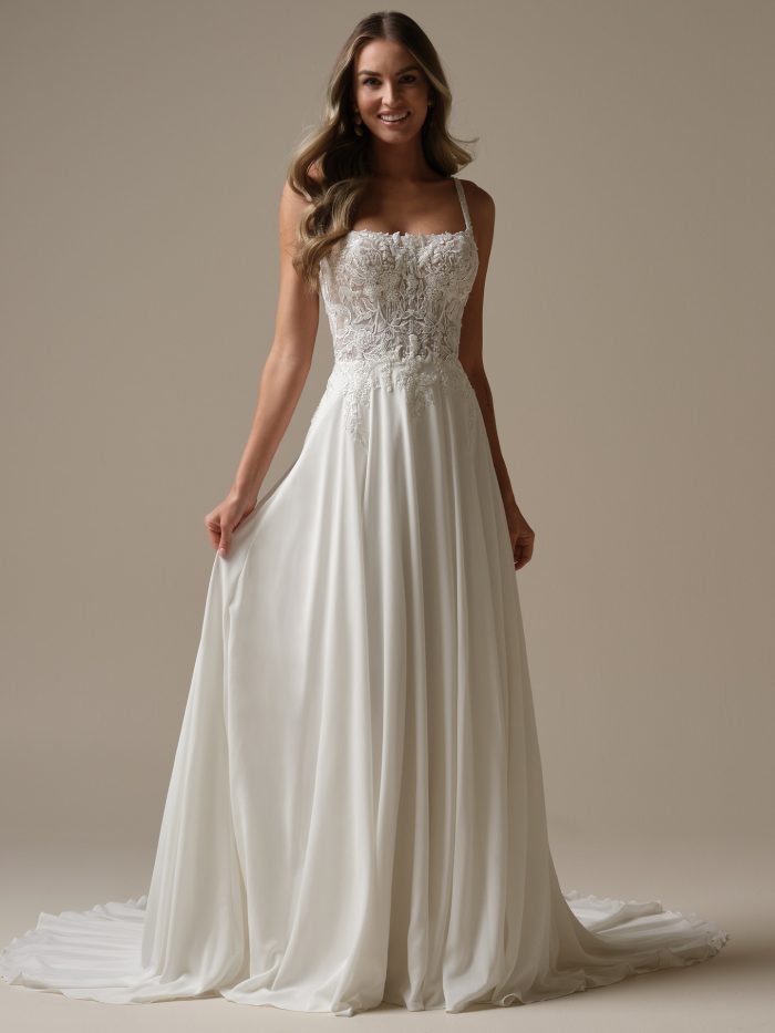 Full-length front view of Avery by Rebecca Ingram an A-line wedding dress with a fitted lace bodice featuring floral appliqués and subtle beadwork, delicate shoulder straps, and a flowing chiffon skirt that drapes to the floor, photographed against a neutral studio background.