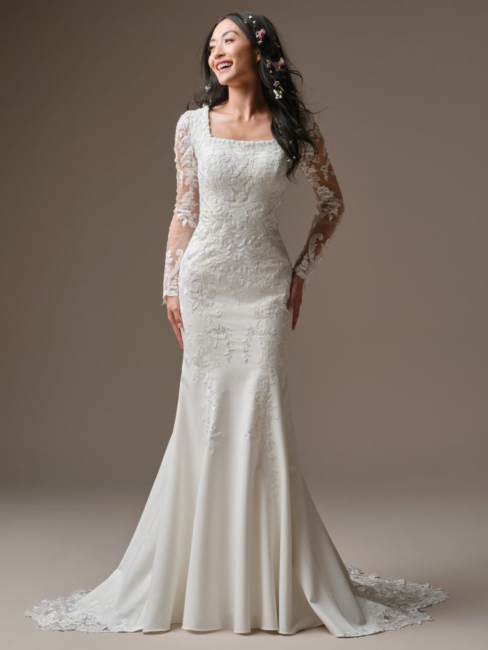 Close-up detail of Larissa Leigh by Rebecca Ingram a sheath wedding dress featuring a fitted lace bodice with floral embroidery, a straight neckline, and long sheer lace sleeves with scalloped edges, photographed against a neutral studio background.