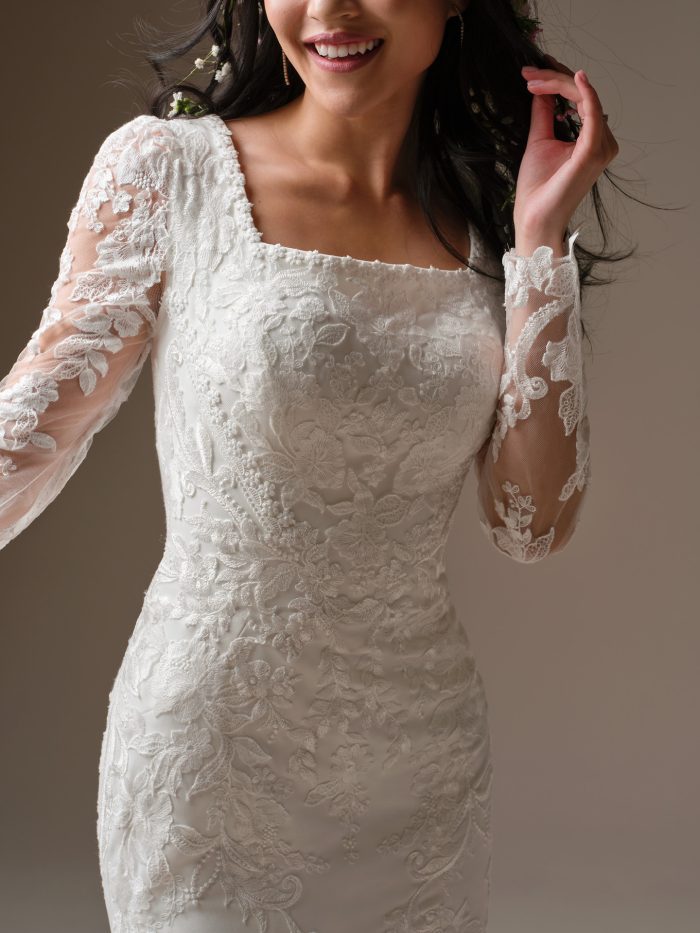 Close-up detail of Larissa Leigh by Rebecca Ingram a sheath wedding dress featuring a fitted lace bodice with floral embroidery, a straight neckline, and long sheer lace sleeves with scalloped edges, photographed against a neutral studio background.