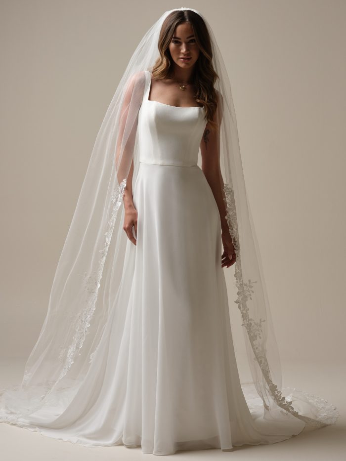 Full-length front view of Olivianna Lane by Rebecca Ingram an A-line wedding dress featuring a clean, structured bodice with a straight neckline and wide shoulder straps, paired with a softly flowing chiffon skirt. The look is styled with a sheer cathedral-length veil trimmed with delicate floral lace along the edges, photographed against a neutral studio background.