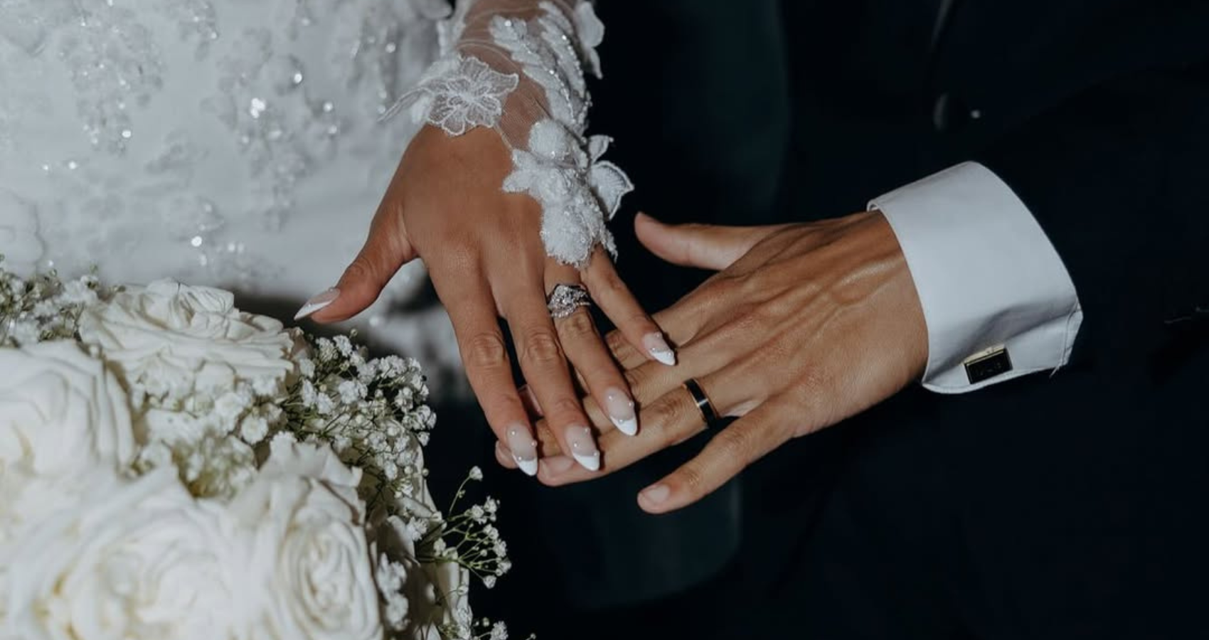 Close-up of two hands wearing wedding rings, one with a lace-embroidered sleeve, resting together beside a bouquet of white roses.