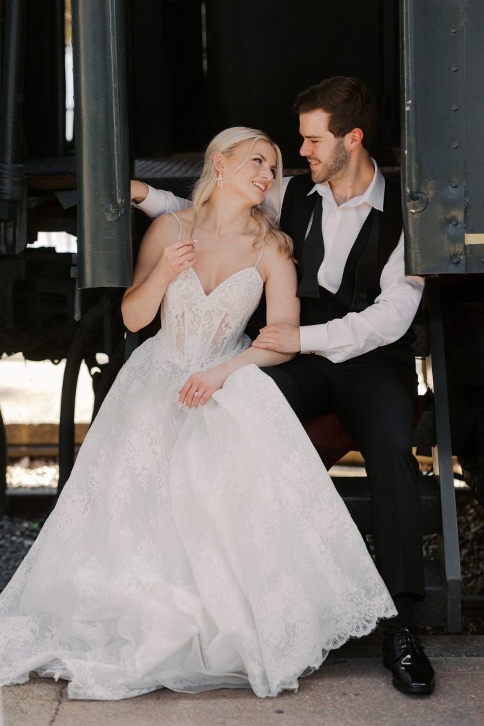 Narissa wedding dress by Sottero and Midgley featuring a lace spaghetti-strap bodice with a soft sweetheart neckline and sheer corset structure. The gown flows into a full lace skirt, styled in a romantic moment with a partner while seated on the steps of a vintage train car.