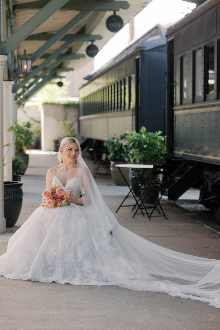 Narissa wedding dress by Sottero and Midgley in a bridal portrait, highlighting the lace spaghetti-strap bodice and voluminous lace skirt. The look is styled with a long trailing veil and bouquet, with the model seated in a wheelchair along a train platform beside a vintage railcar.