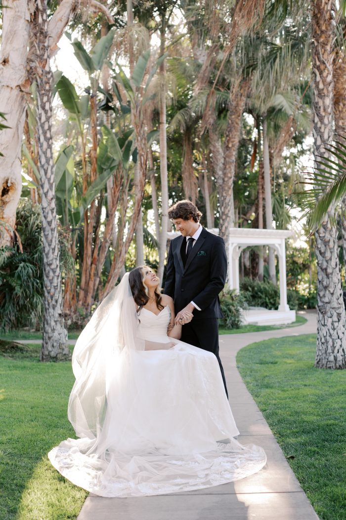 Person seated in a wheelchair on a garden pathway lined with palm trees, holding hands with another individual while wearing the Ethelinda gown by Rebecca Ingram—a strapless, flowing tulle wedding dress with a long train and veil—set against lush greenery and a white gazebo.