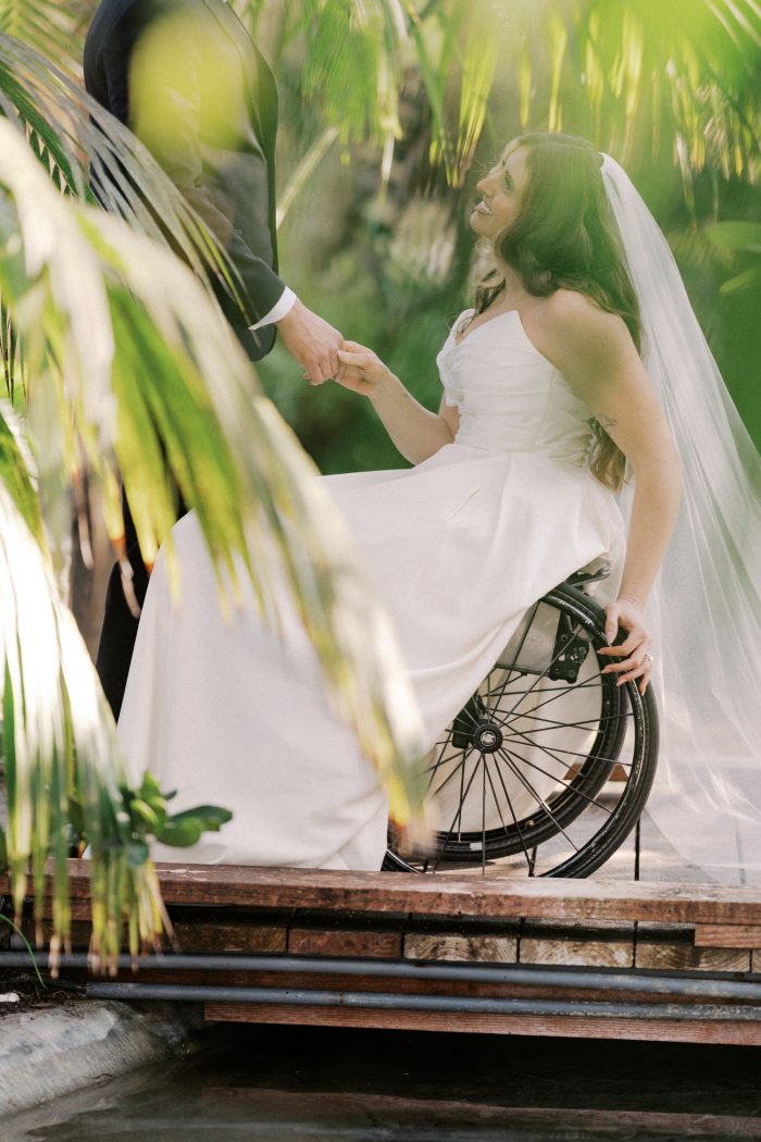 Ethelinda wedding dress by Rebecca Ingram in a lifestyle moment, highlighting the strapless draped bodice and voluminous satin skirt. The gown is styled with a veil while seated in a wheelchair, framed by greenery as a partner’s hand reaches in.