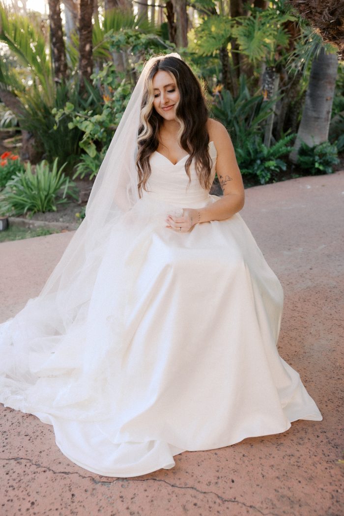 Ethelinda wedding dress by Rebecca Ingram featuring a strapless sweetheart neckline with softly draped bodice and a full ballgown skirt in smooth satin. The look is styled with a long veil in a lush outdoor garden setting.