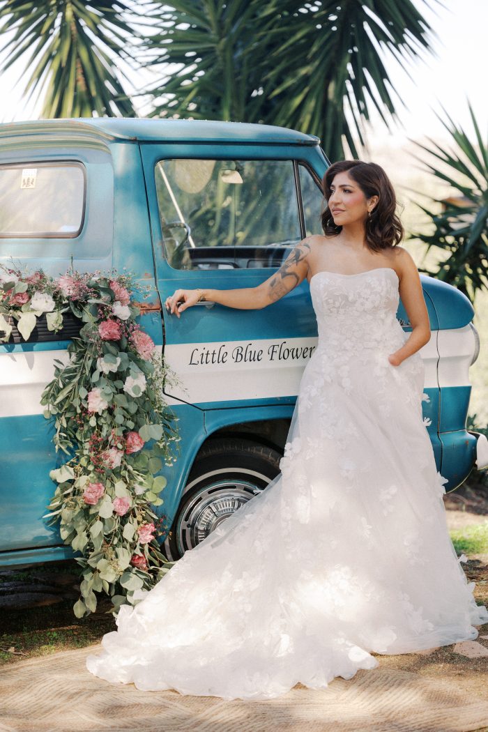 A bride wearing the Laila gown by Maggie Sottero stands beside a vintage blue "Little Blue Flower" truck beneath palm trees, gazing elegantly to the side. The strapless A-line gown showcases an allover 3D floral appliqué design from the structured bodice through the sweeping layered tulle skirt and train. A cascading floral arrangement of pink peonies and eucalyptus decorates the truck door beside her.