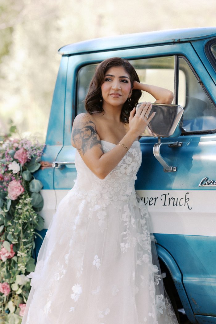 A bride wearing the Laila gown by Maggie Sottero leans against a vintage blue flower truck adorned with a lush arrangement of pink roses and eucalyptus. The strapless A-line gown features an allover 3D floral appliqué bodice and a voluminous layered tulle skirt. She accessorizes with gold hoop earrings and a bracelet, and her dark wavy hair is swept to one side.