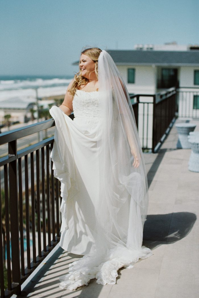 A bride wearing the Avery gown by Rebecca Ingram stands on a rooftop terrace overlooking the ocean, laughing joyfully. Her strapless lace-bodice gown features a flowing skirt with a lace-trimmed cathedral train, paired with a long sheer veil that catches the breeze. Her blonde hair falls in soft waves over one shoulder.