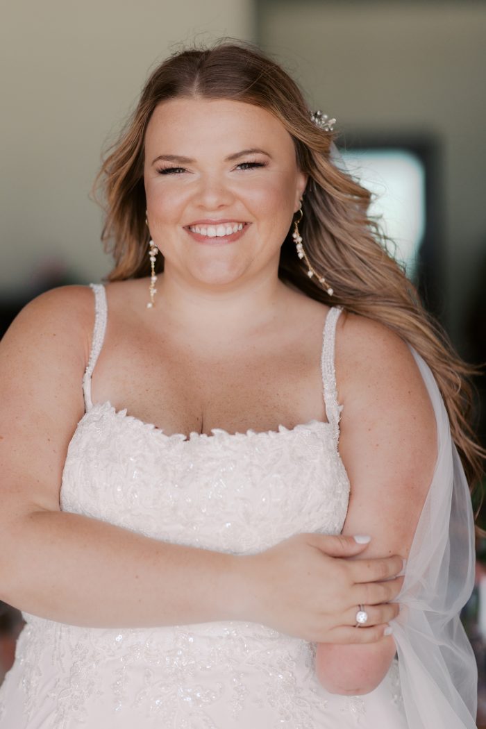 A close-up portrait of a bride wearing the Avery gown by Rebecca Ingram, smiling warmly at the camera with her arms crossed. The spaghetti-strap, square-neck bodice features delicate floral lace and subtle sequin detailing. She accessorizes with dangling pearl and gold earrings, a small floral hair pin, and a round-cut solitaire engagement ring, with a sheer veil draped over her shoulder.