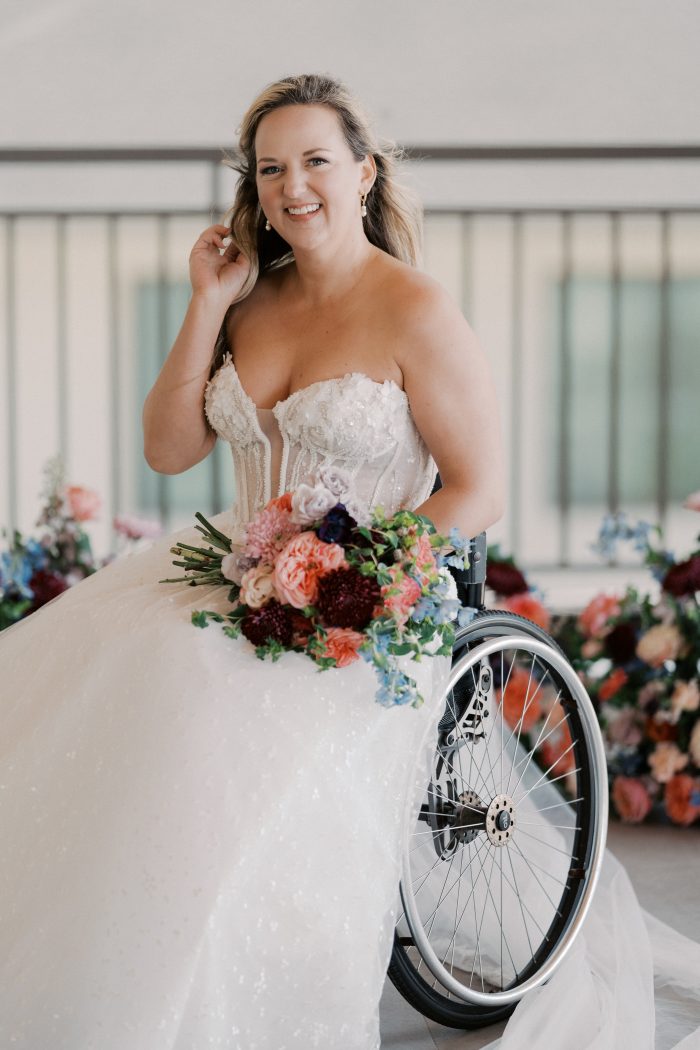 Athlone wedding dress by Sottero and Midgley featuring a strapless sweetheart neckline with dimensional floral appliqués and a sheer corset bodice. The gown flows into a full, softly textured skirt, styled with a colorful bouquet, with the model seated in a wheelchair in a bright indoor setting.