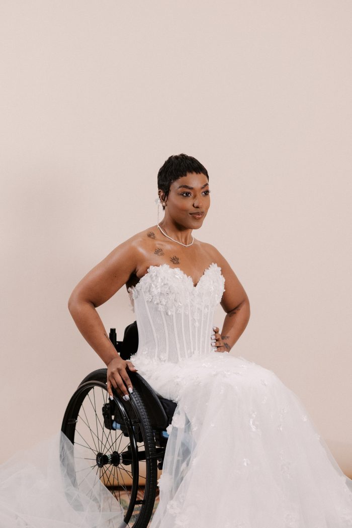 Akari wedding dress by Sottero and Midgley featuring a strapless sweetheart neckline with dimensional floral appliqués over a structured corset bodice. The gown flows into a voluminous tulle skirt with soft floral detailing, styled seated in a wheelchair against a neutral studio backdrop.