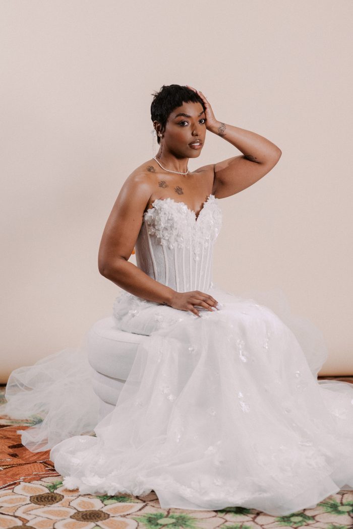 Akari wedding dress by Sottero and Midgley in a seated studio portrait, highlighting the strapless floral-appliqué bodice with sheer corsetry, and a full airy tulle skirt with scattered three-dimensional floral accents.