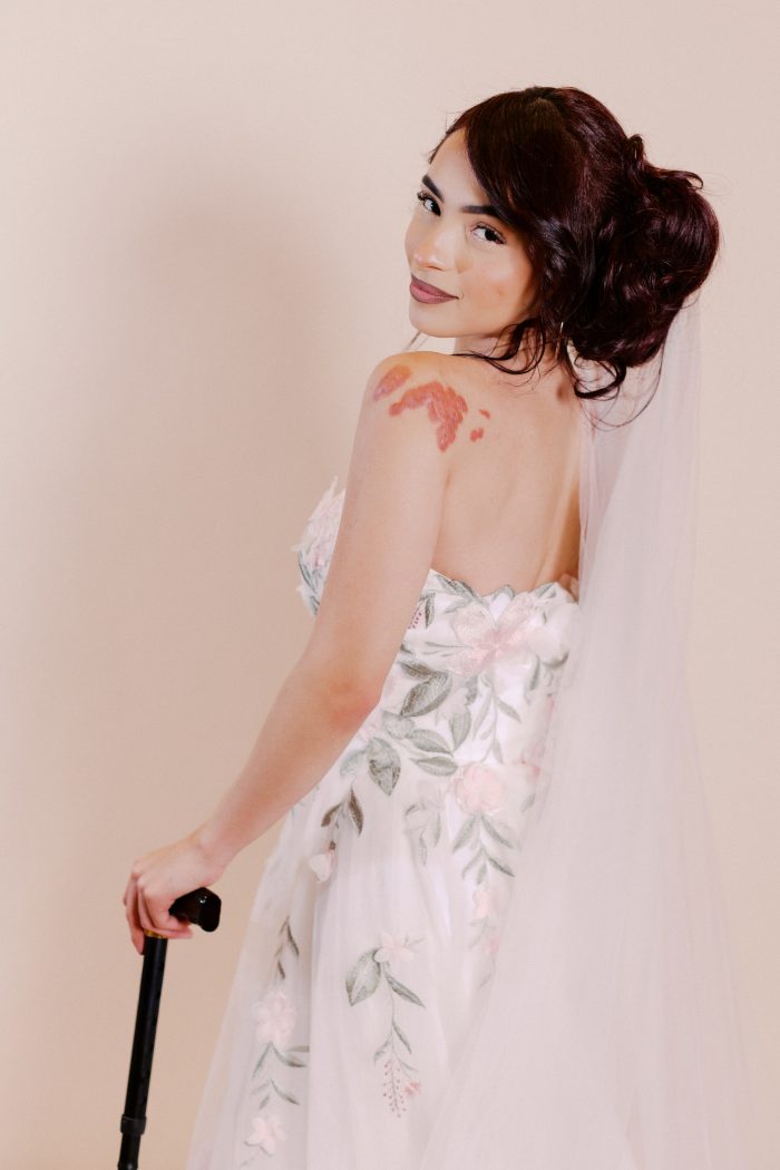 Blossom wedding dress by Rebecca Ingram in a close-up back view, highlighting the strapless floral-appliqué bodice, soft tulle layers, and coordinating embroidered details. The look is styled with a veil, with the model holding a cane, showcasing the gown’s romantic, garden-inspired design.